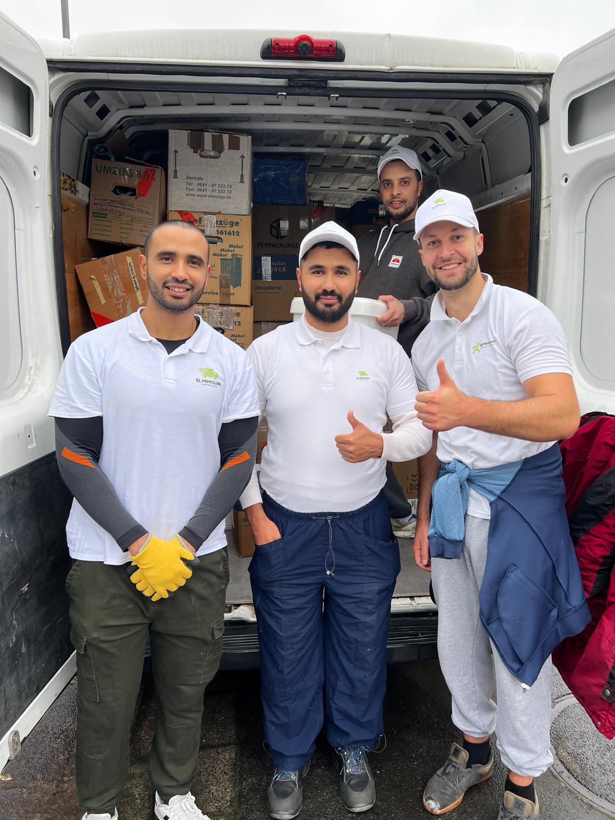 Four men posing at the back of a delivery van filled with packed boxes, three wearing white El Mimouni polo shirts and caps, one giving thumbs up.