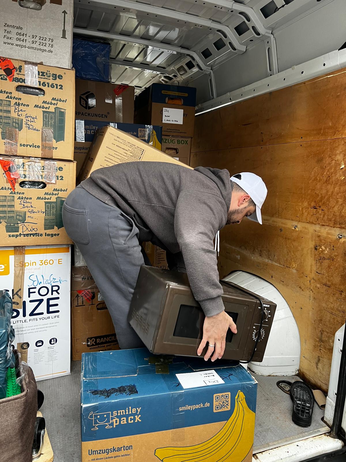 Man wearing a white cap and gray hoodie lifting a microwave inside a moving van filled with packed boxes.