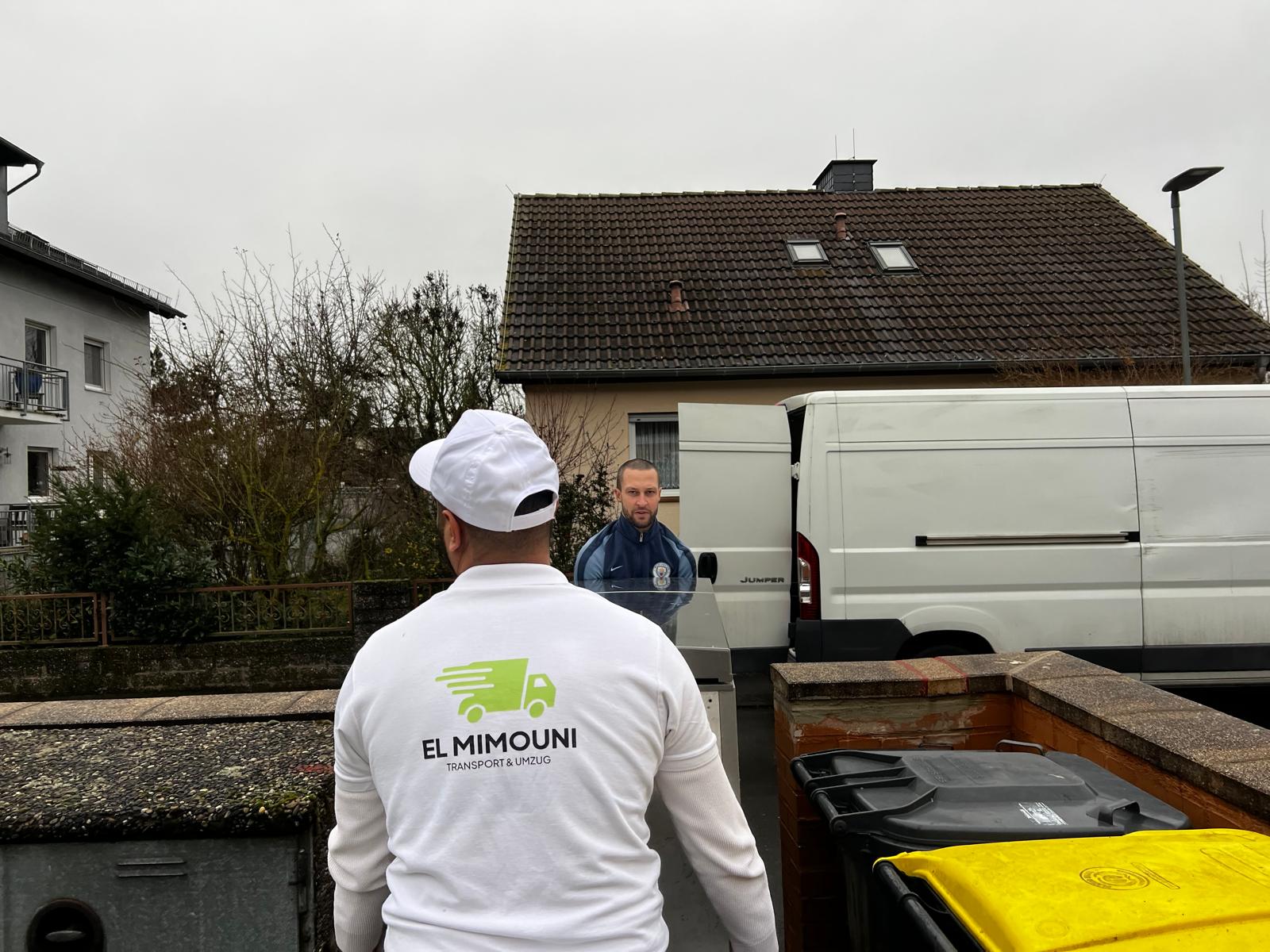 Two men near a white van, one wearing a white shirt and cap with 'EL MIMOUNI TRANSPORT & UMZUG' logo on the back.