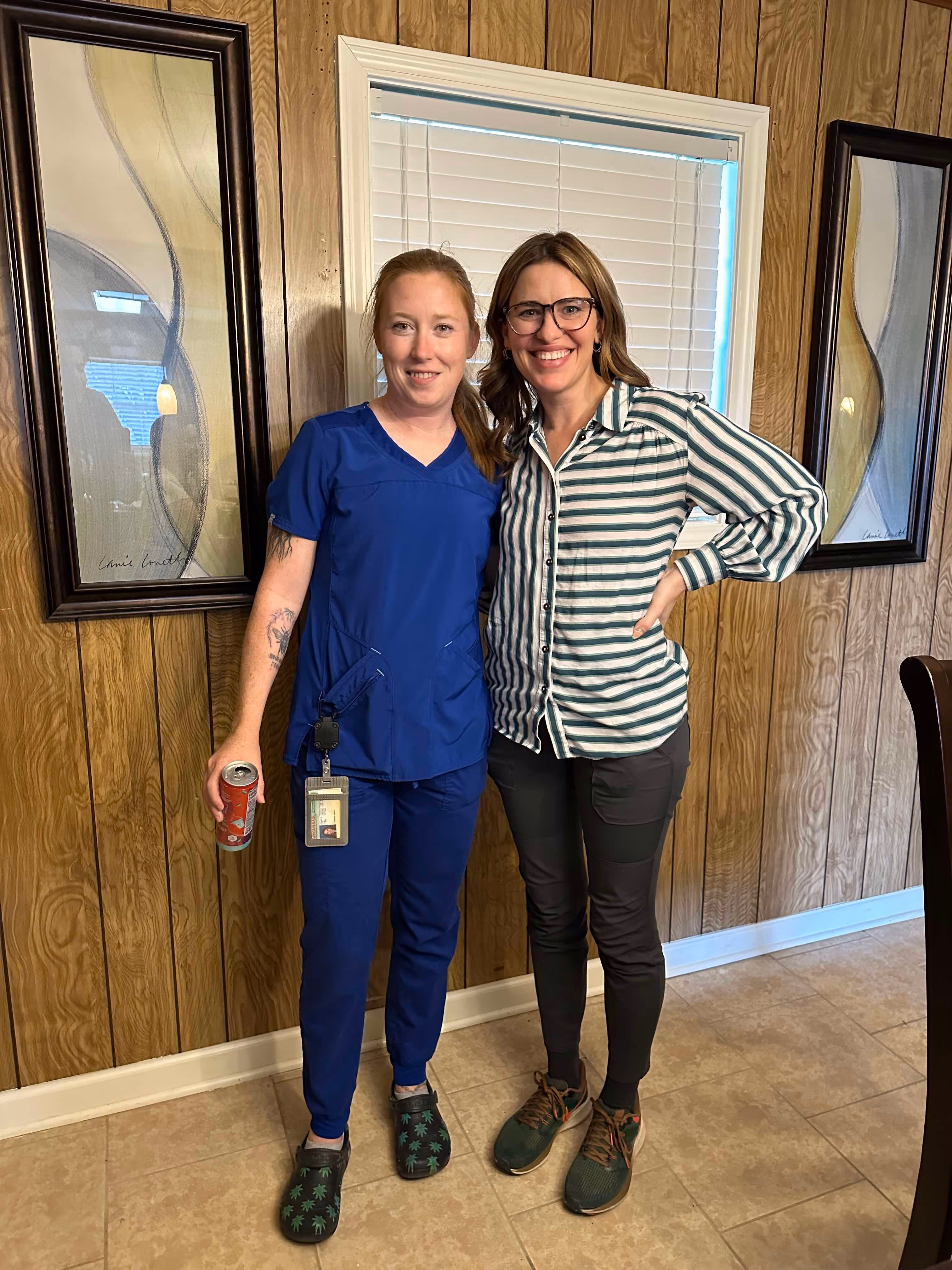 One woman wearing blue scrubs with red hair hugs another woman with green striped shirt in a room with wood paneling