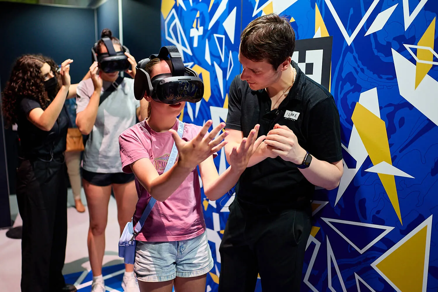 A man guides a girl wearing a VR headset who is moving her hands, while other people in the background also wear VR headsets.
