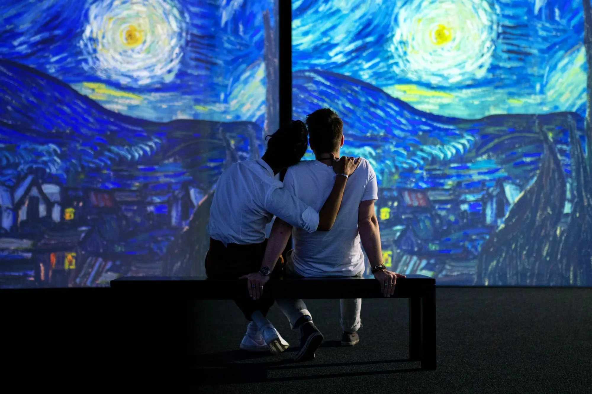 Two people sitting closely on a bench, one resting their head on the other's shoulder, in front of a large illuminated display of Van Gogh's Starry Night.