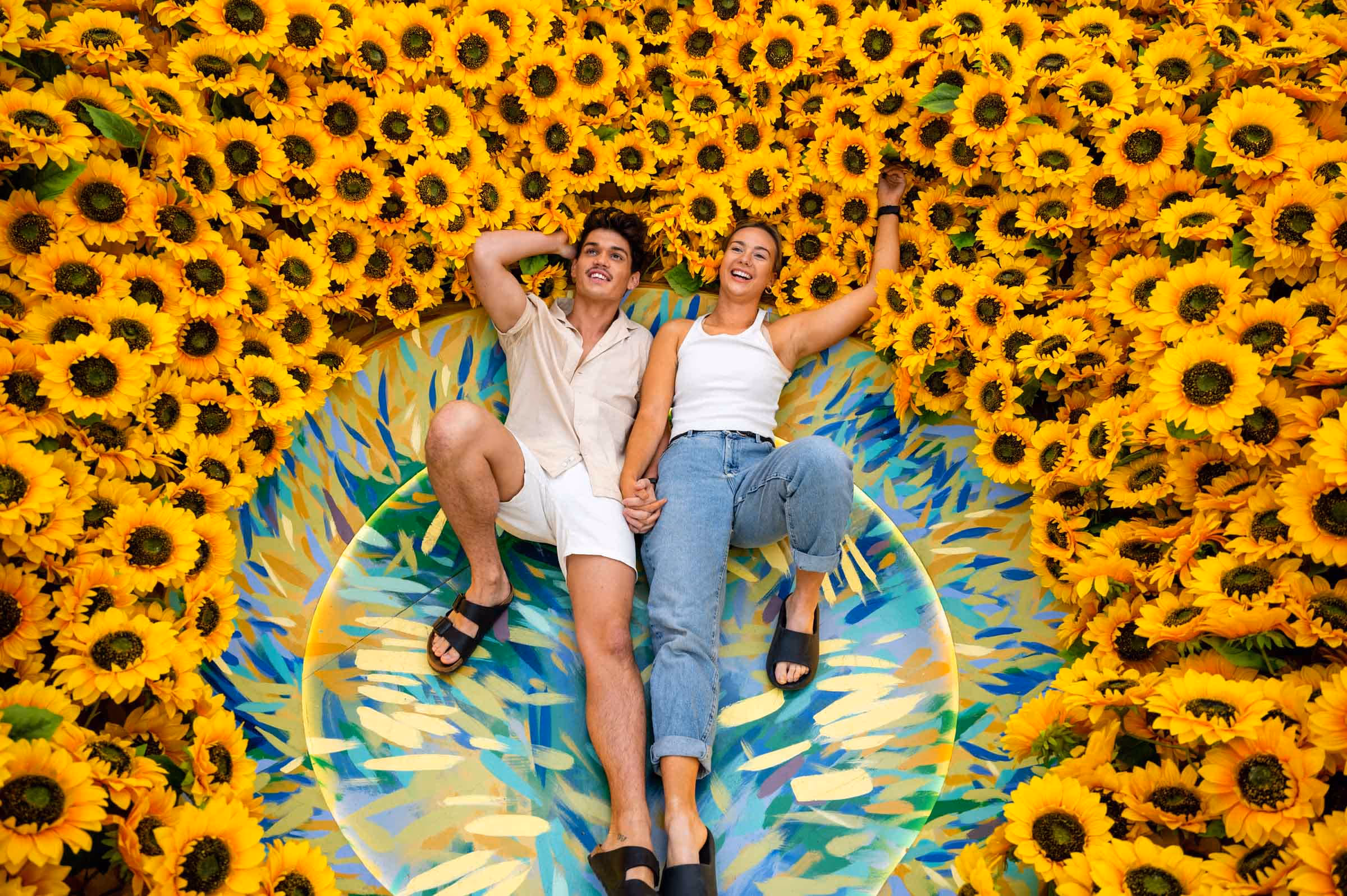 A smiling couple lies holding hands on a colorful patterned circular surface surrounded by vibrant yellow sunflowers.