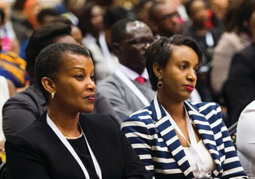 Two women seated among an audience, attentively listening at an event.