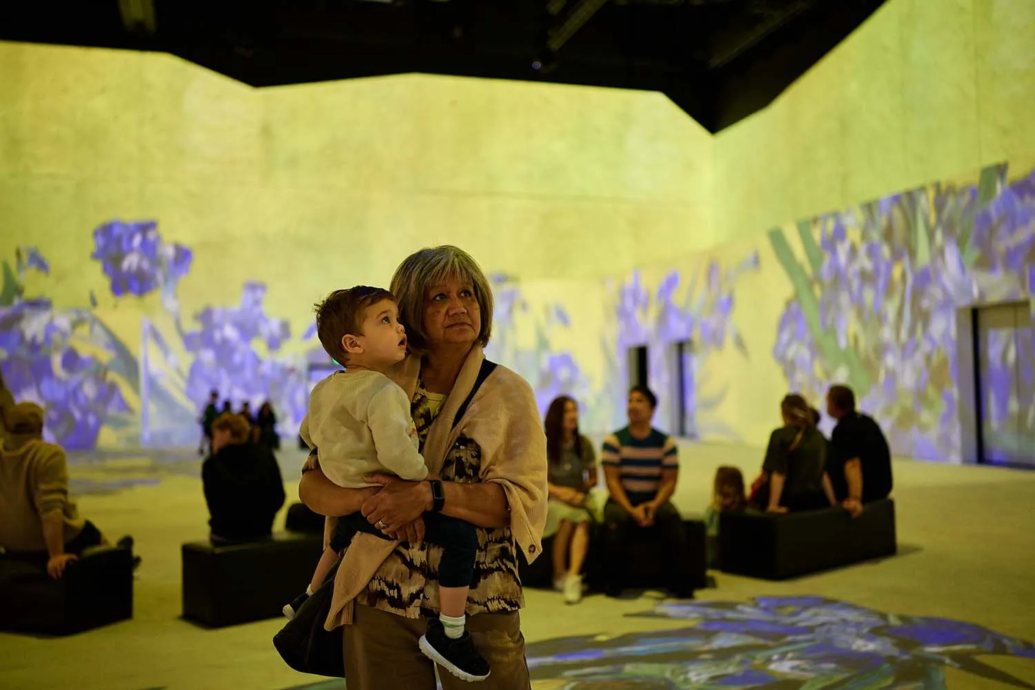 Older woman holding a young boy inside a dimly lit room with large walls showing a purple floral projection, other people seated in the background.