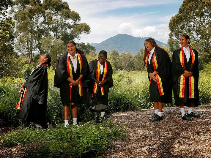 Five young women in black graduation gowns with red and yellow stoles standing and laughing outdoors with trees and hills in the background.
