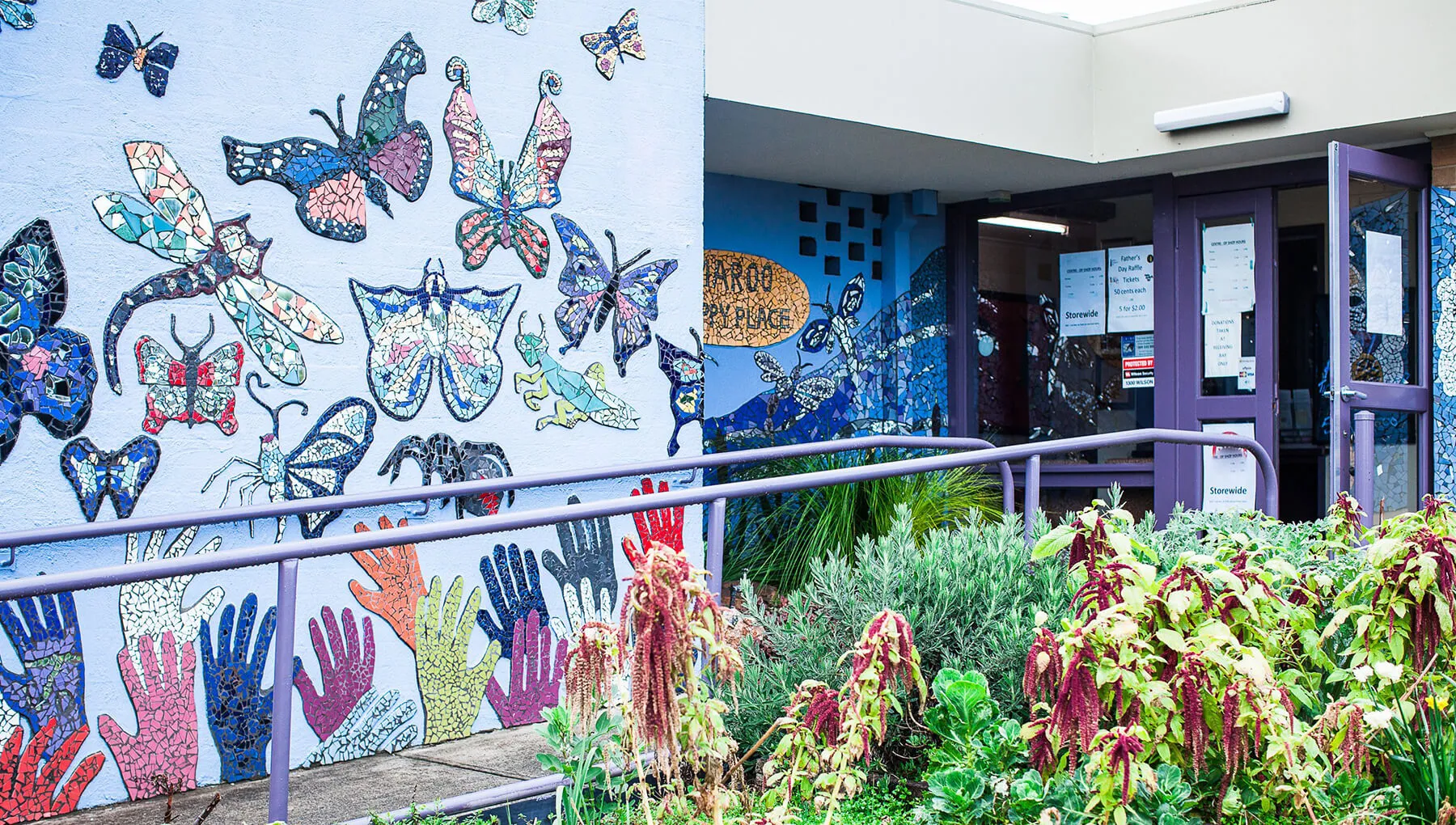 Entrance to a building with a purple door and railing, decorated with a colorful mosaic mural of butterflies, dragonflies, and handprints on the walls.