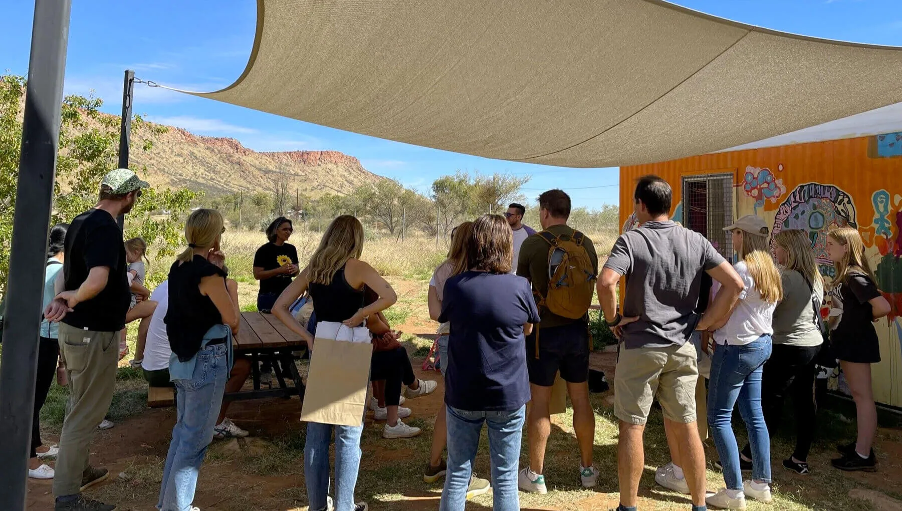 Group of people gathered outdoors under a shade sail near a painted orange structure with mountains in the background.