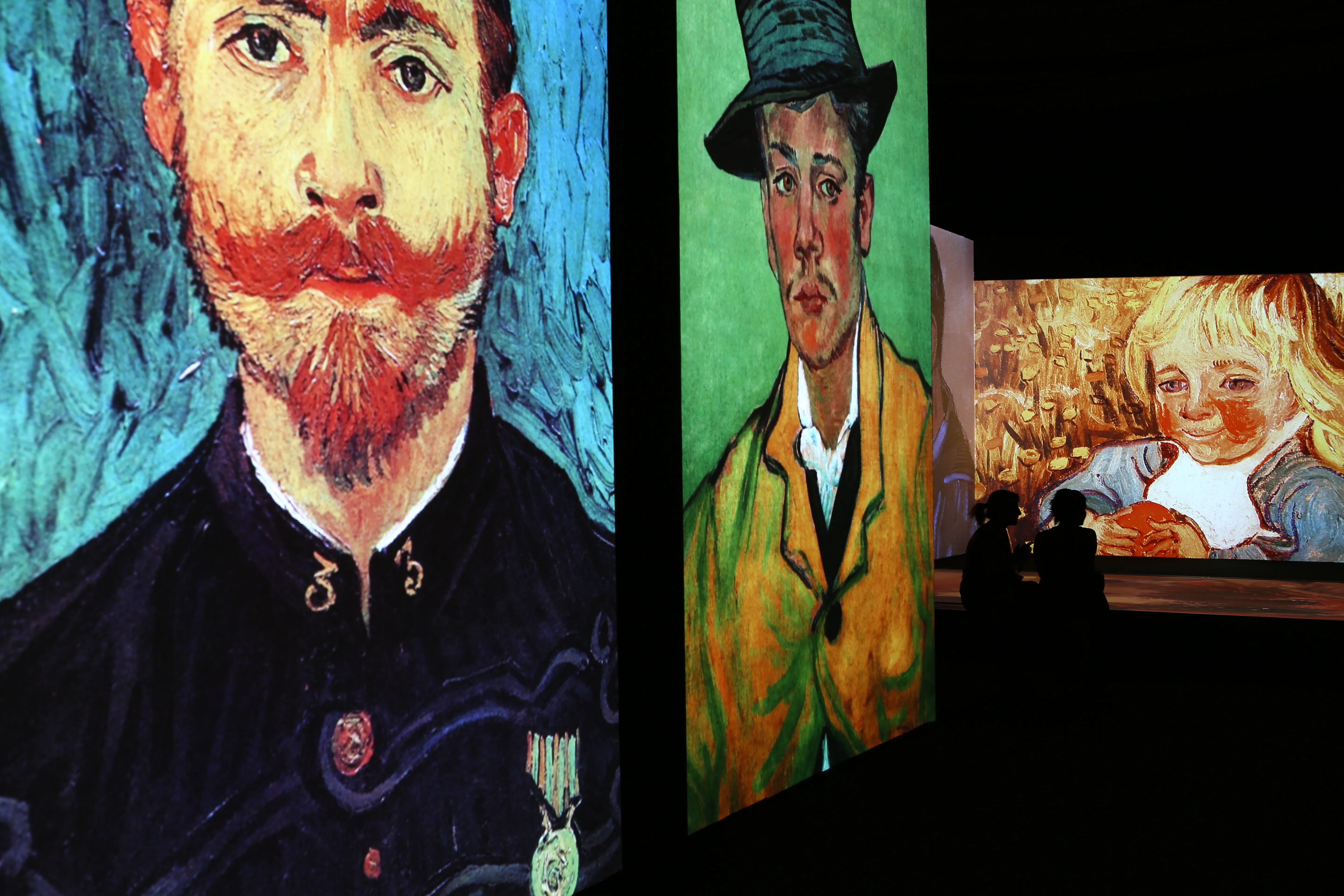 Indoor art exhibition with large, vertical projections of classic paintings including a bearded man, a man in a hat, and a blonde child, with two silhouetted visitors seated in front.