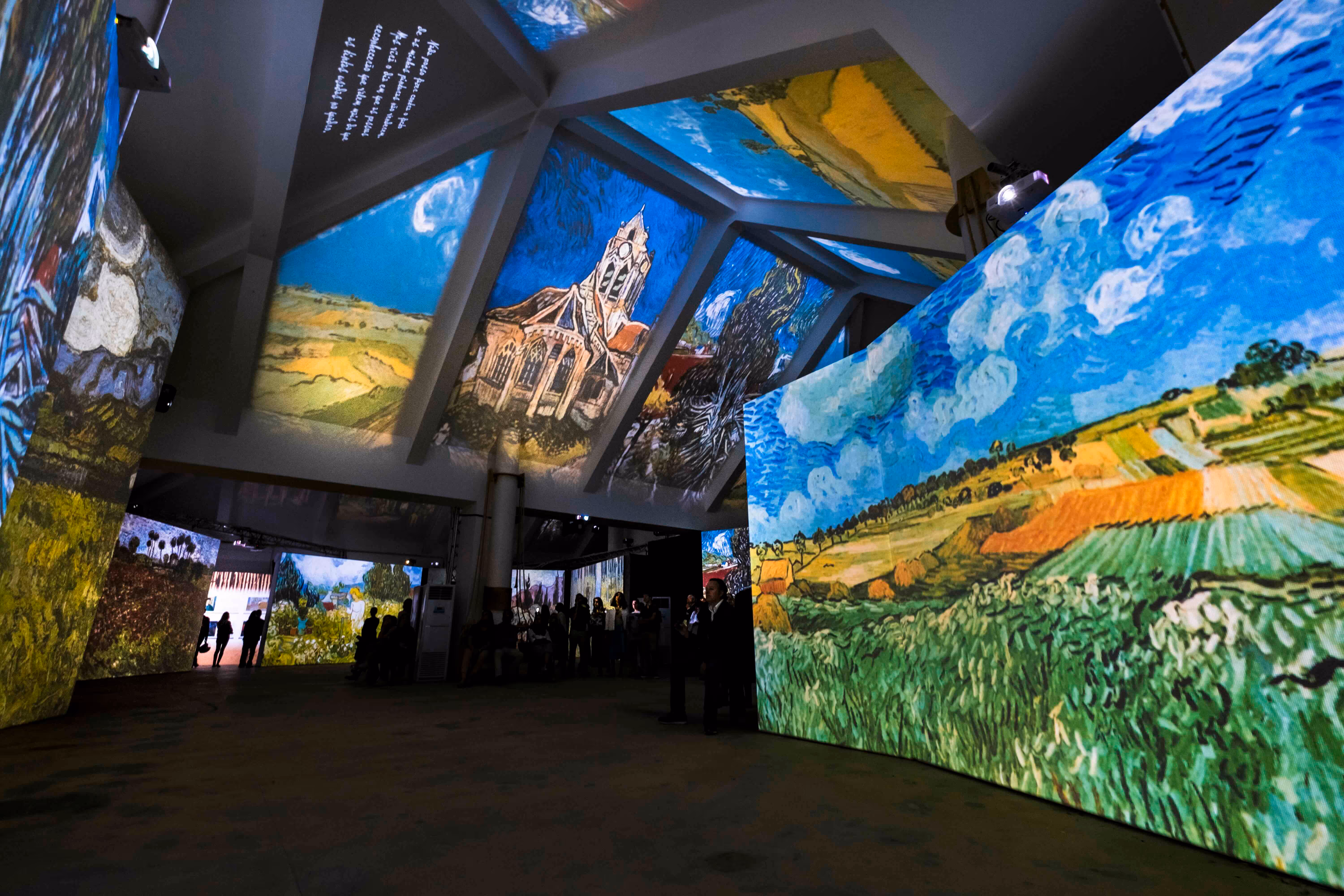 Indoor exhibition of large digital projections featuring Van Gogh-style paintings of landscapes and buildings with people observing.