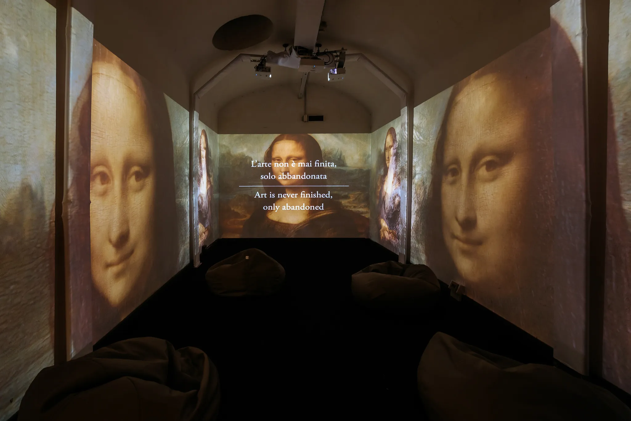 Projection of Mona Lisa images on multiple walls with quote about art displayed in a dark room with bean bags.