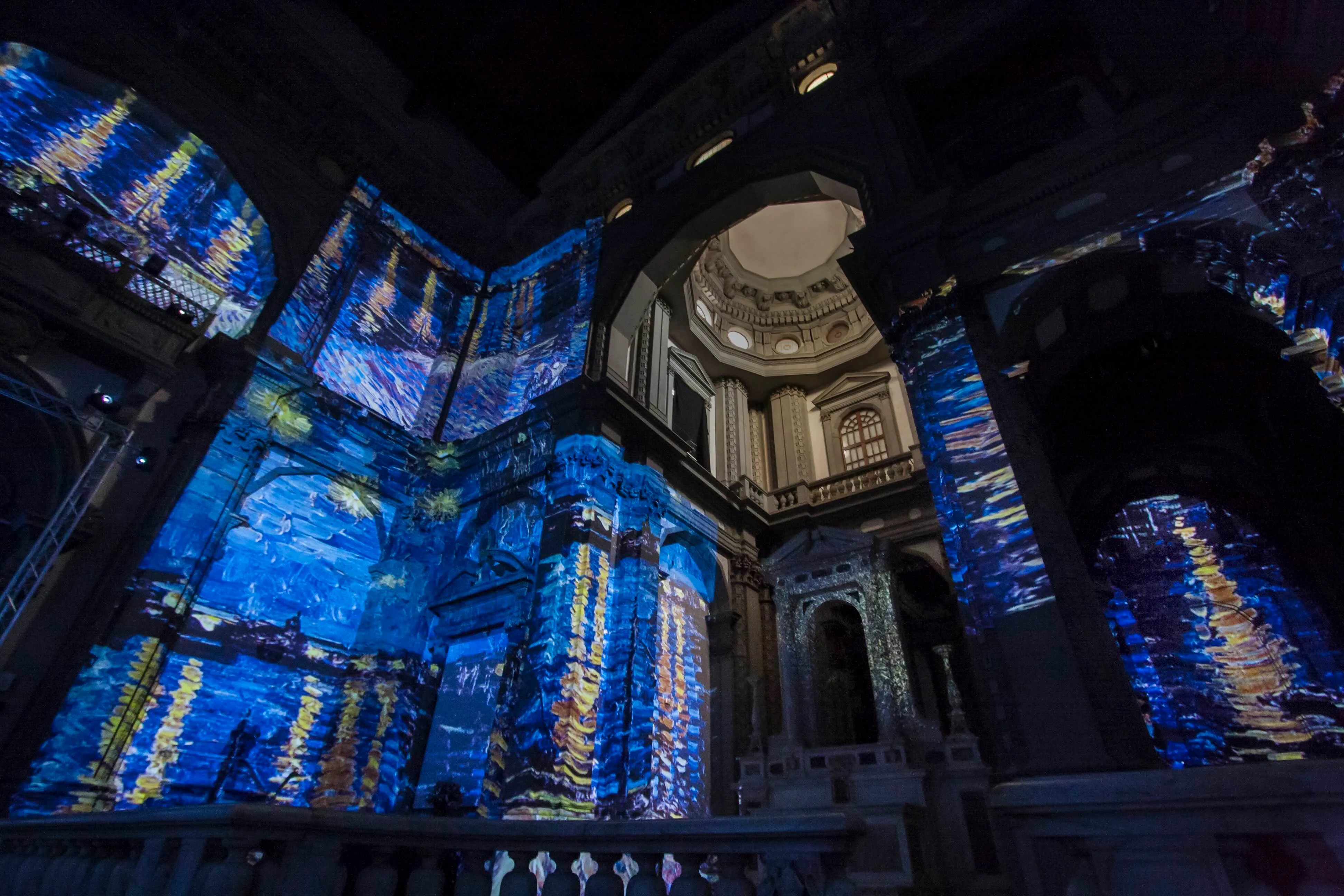 Interior of a historic building with walls and arches illuminated by blue and yellow projections resembling starry night reflections.