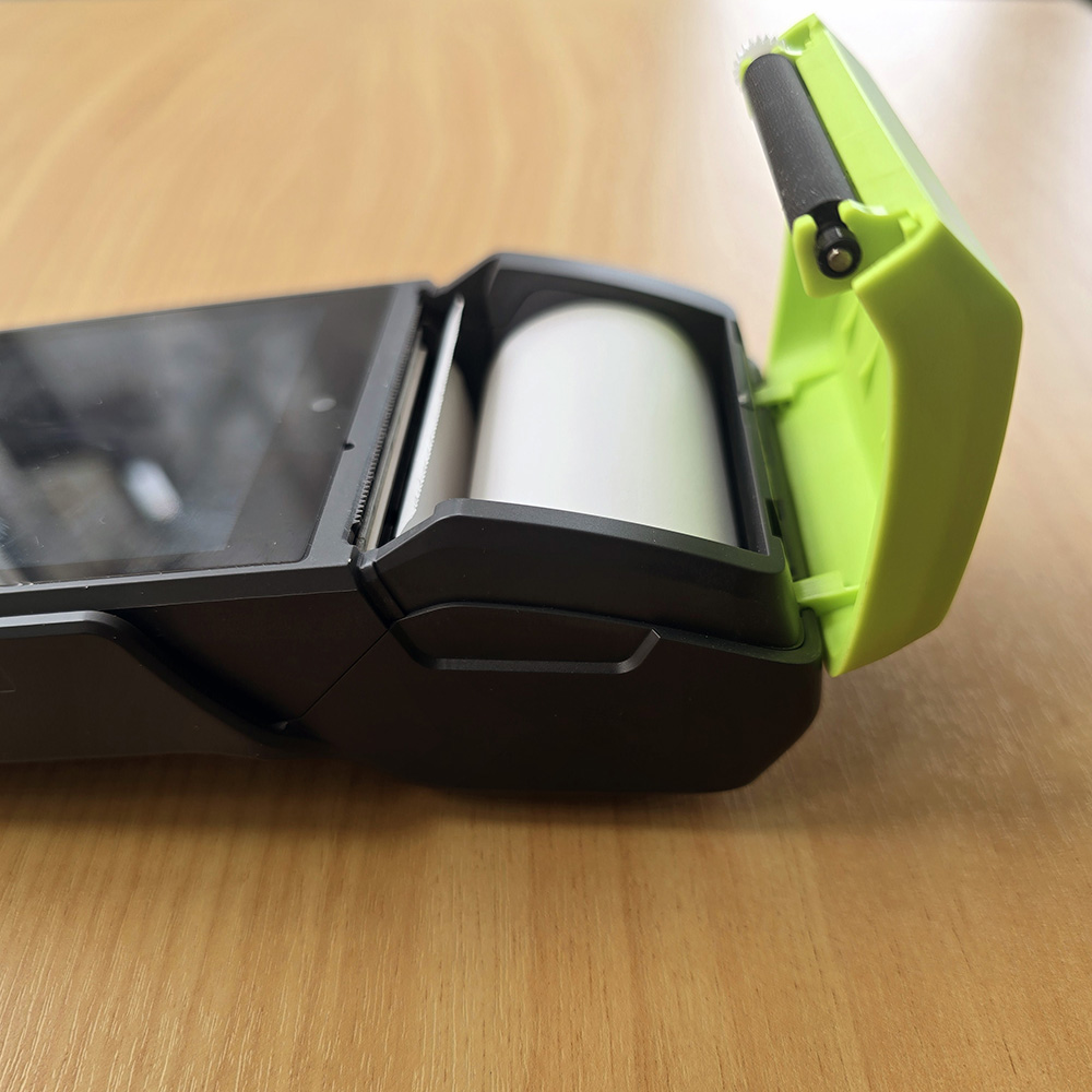 Portable handheld receipt printer with its green cover open, showing the paper roll inside.
