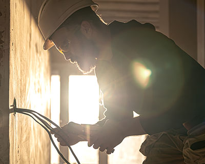 Man in a hard hat using pliers or wire cutters on a wall with sunlight behind him.