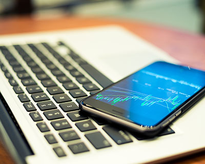 Smartphone showing financial charts resting on a laptop keyboard on a wooden desk.