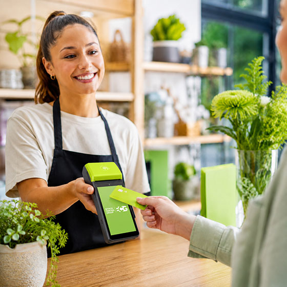 Smiling cashier in apron holding card reader as customer taps a green card to pay at flower shop.