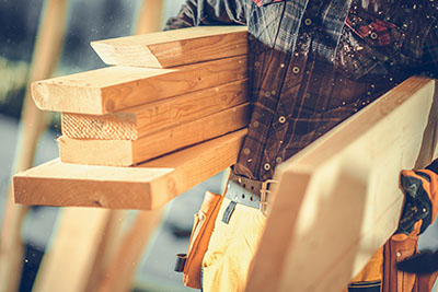 Construction worker wearing a plaid shirt and yellow pants carrying several pieces of lumber at a building site.