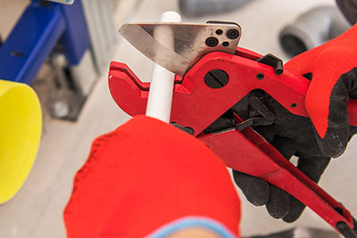 Hands wearing red gloves cutting a white PVC pipe using red pipe cutters.