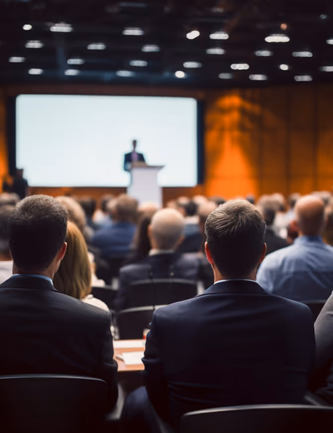 Das Publikum sitzt in einem Konferenzsaal und hört einem Redner zu, der auf der Bühne vor einer großen Leinwand spricht.