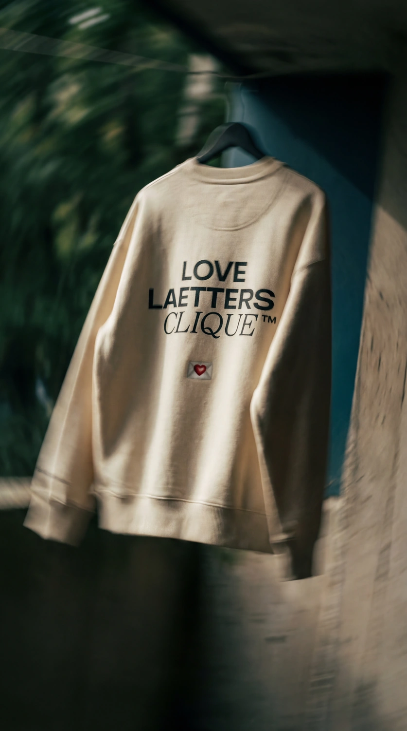 Beiger Pullover mit der Aufschrift „LOVE LAETTERS CLIQUE™“ und einem kleinen Brief mit rotem Herz darunter.