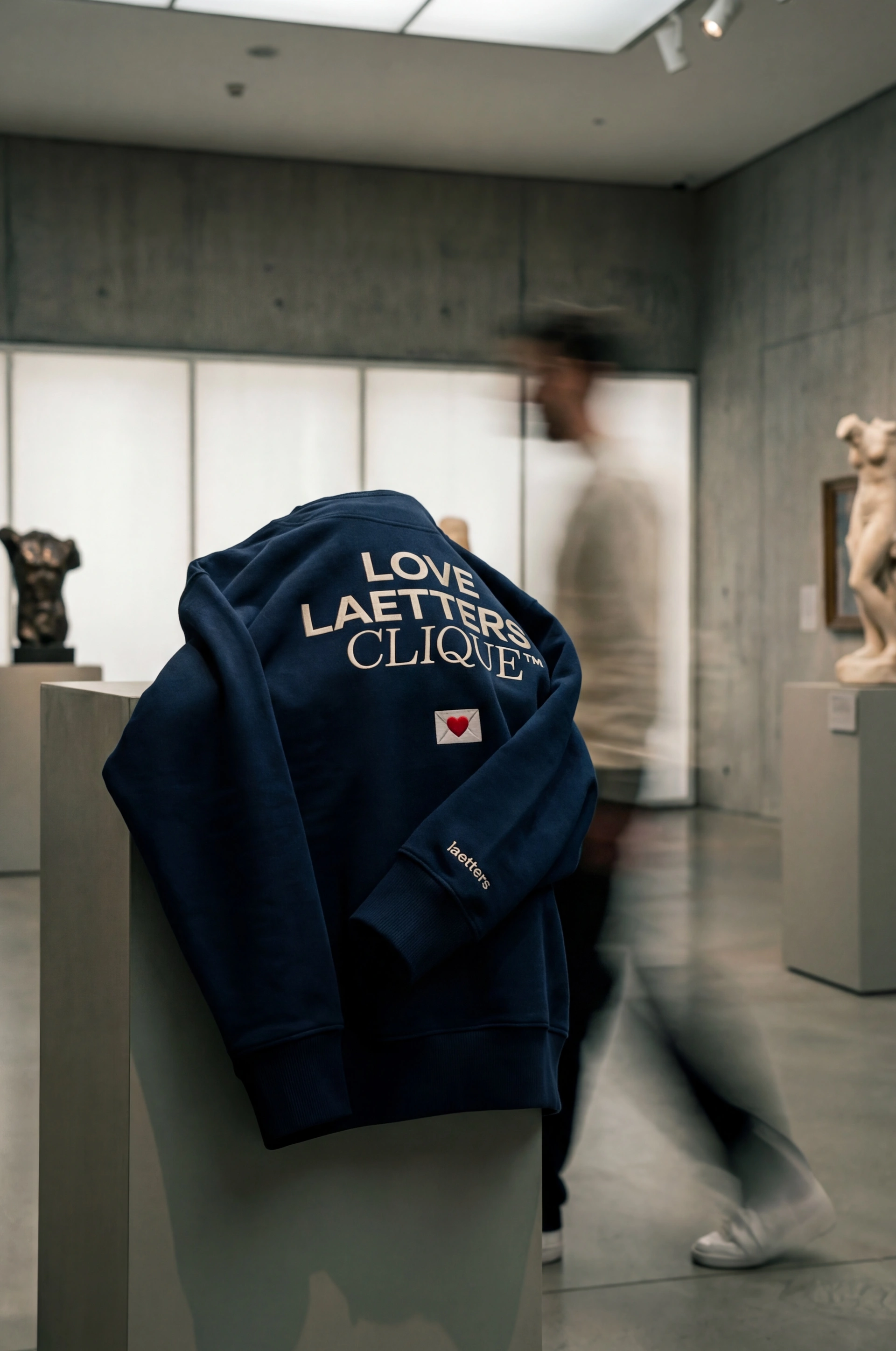 Dunkelblauer Pullover mit der Aufschrift "LOVE LAETTERS CLIQUE" hängt auf einem weißen Kleiderständer in einem modernen Ausstellungsraum mit Betonwänden und großen Fenstern, unscharfe Personen gehen vorbei.