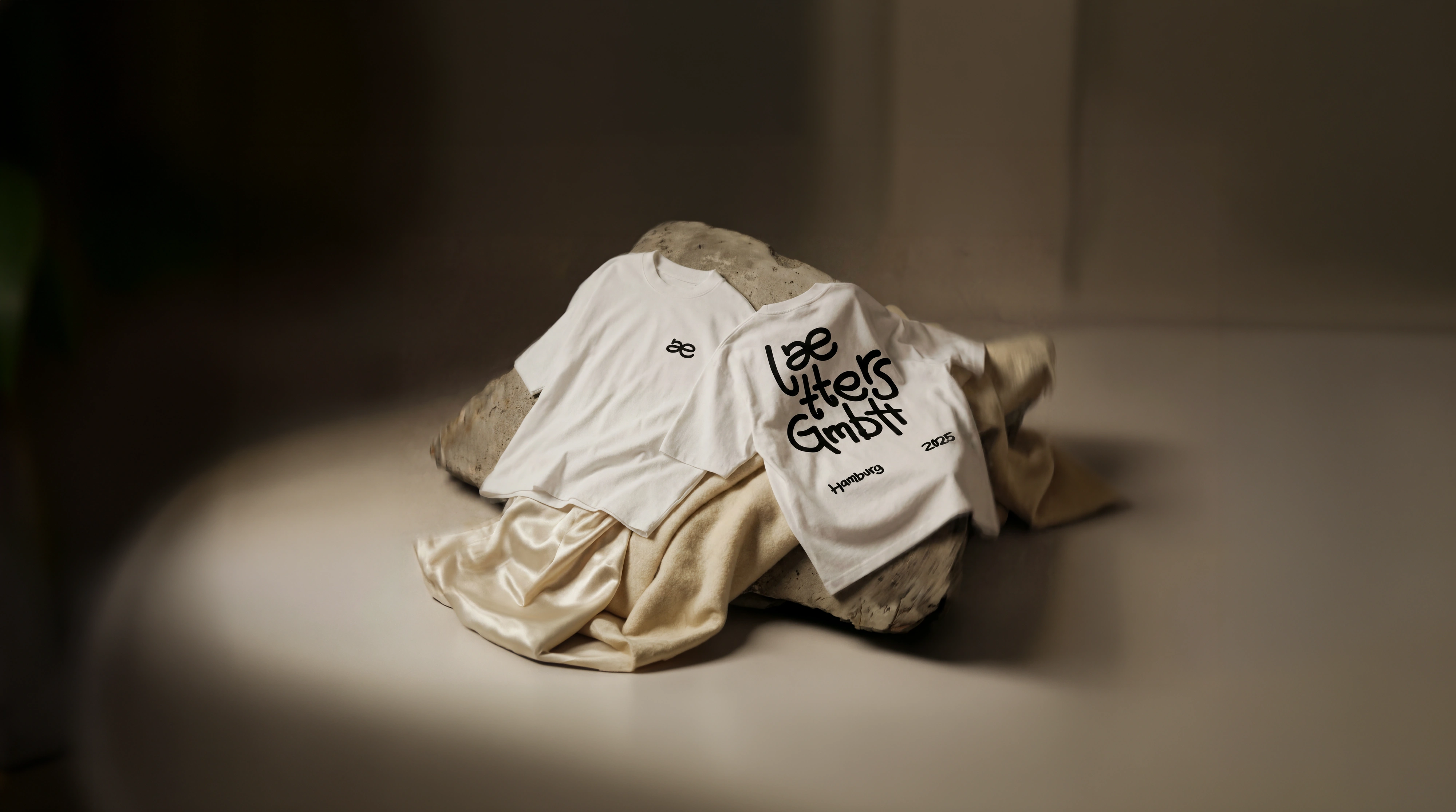 Zwei weiße laetters couture T-Shirts mit schwarzem Aufdruck liegen auf einem mit Stoff bedeckten Stein.