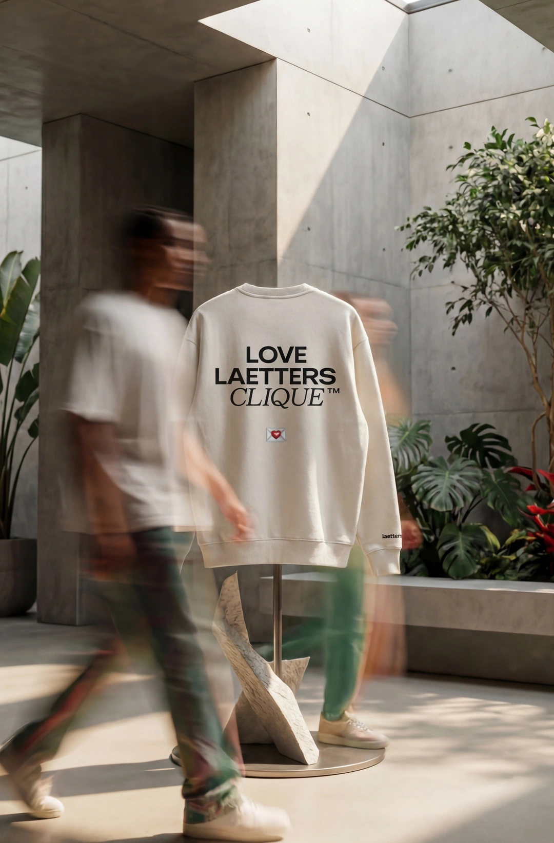 Weißes T-Shirt mit Aufschrift 'Love letters no e-mails' und einem Herz auf einem Kleiderständer in einem modernen Innenraum mit Pflanzen, davor verschwommene gestreifte Menschen.