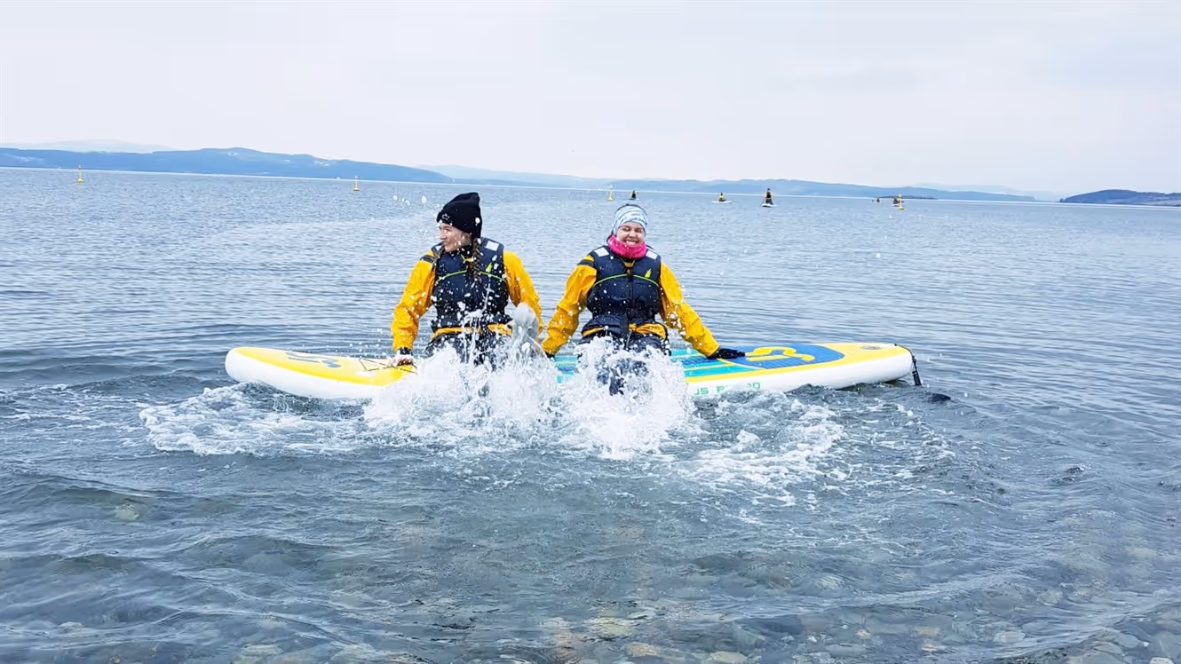 To personer i vannsikre jakker sitter på et stand-up paddleboard og plasker bena i klart vann, med et landskap i det fjerne og et smil om munnen.
