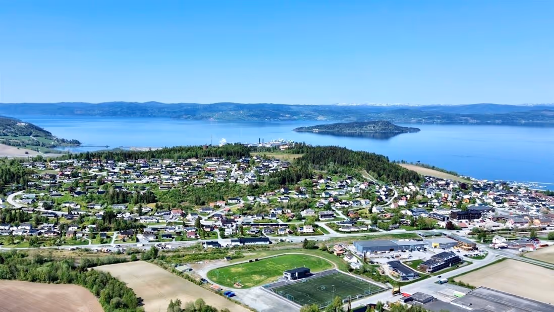 "Luftfoto av en liten by ved sjøen med tett boligbebyggelse og frodig vegetasjon, omgitt av en blå fjord.