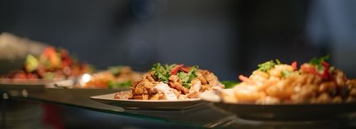 Serveringsfat med et utvalg av mat, fokusert på en tallerken med grillet kylling toppet med urter, med andre retter i uskarphet i bakgrunnen.