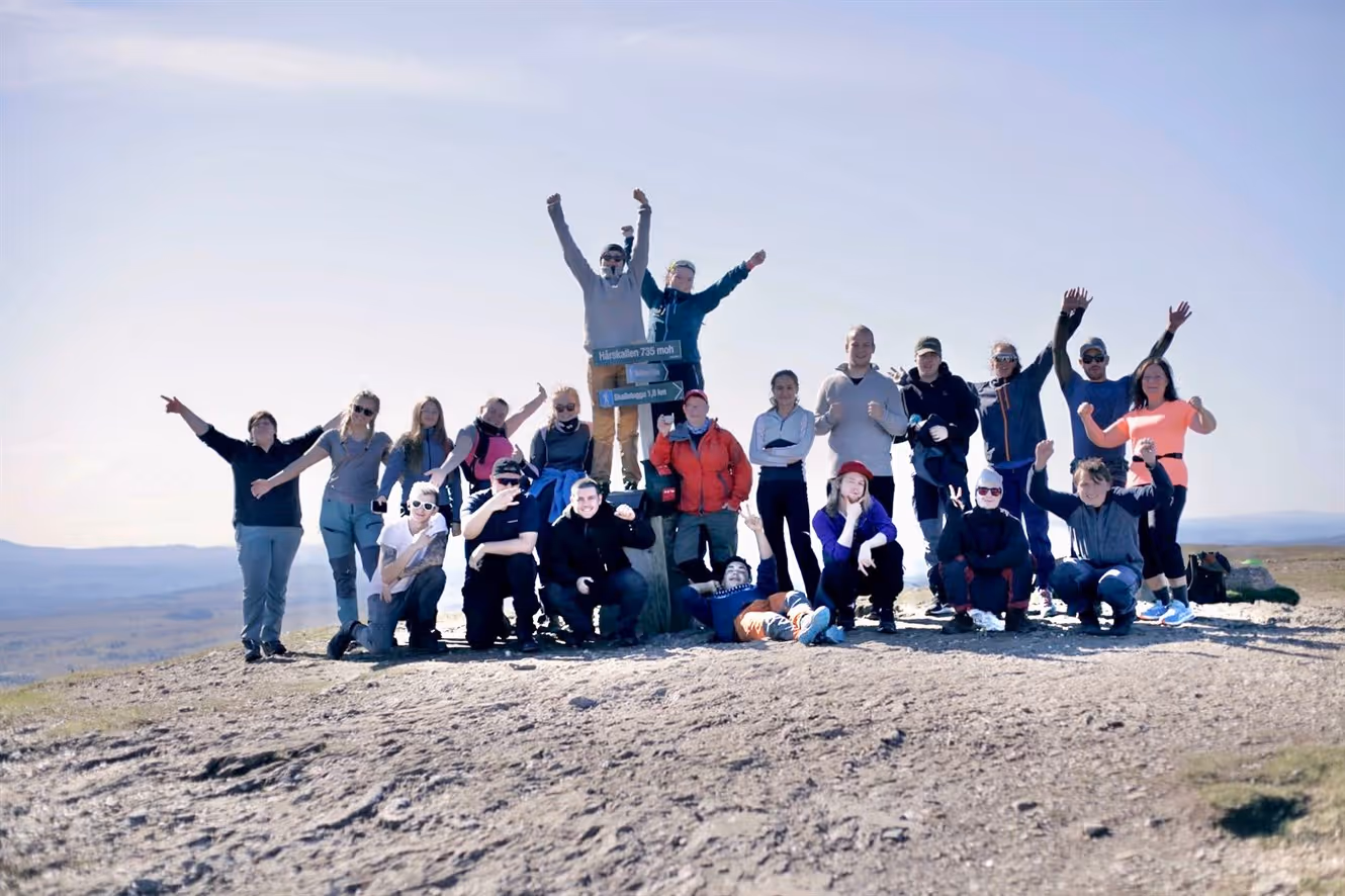 En gruppe elever og lærere feirer på toppen av et fjell med klar himmel, noen står, andre sitter, og flere løfter armene i triumf.