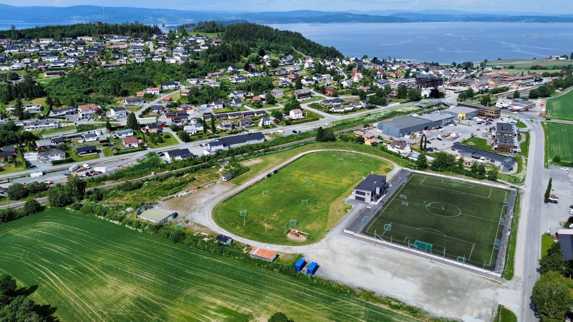 Luftbilde av tettstedet Skogn med bolighus, et fotballfelt og en lekeplass, med fjell og en fjord i bakgrunnen.