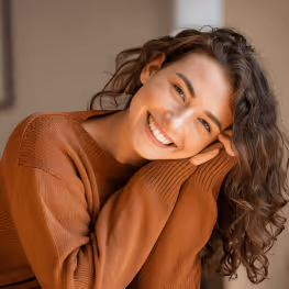 Smiling woman with curly hair resting her head on her hands wearing a brown sweater.