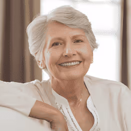 Smiling elderly woman with short white hair wearing a white blouse, sitting indoors.