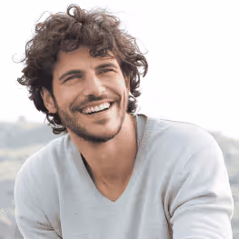 Smiling man with curly hair wearing a light gray sweater outdoors.