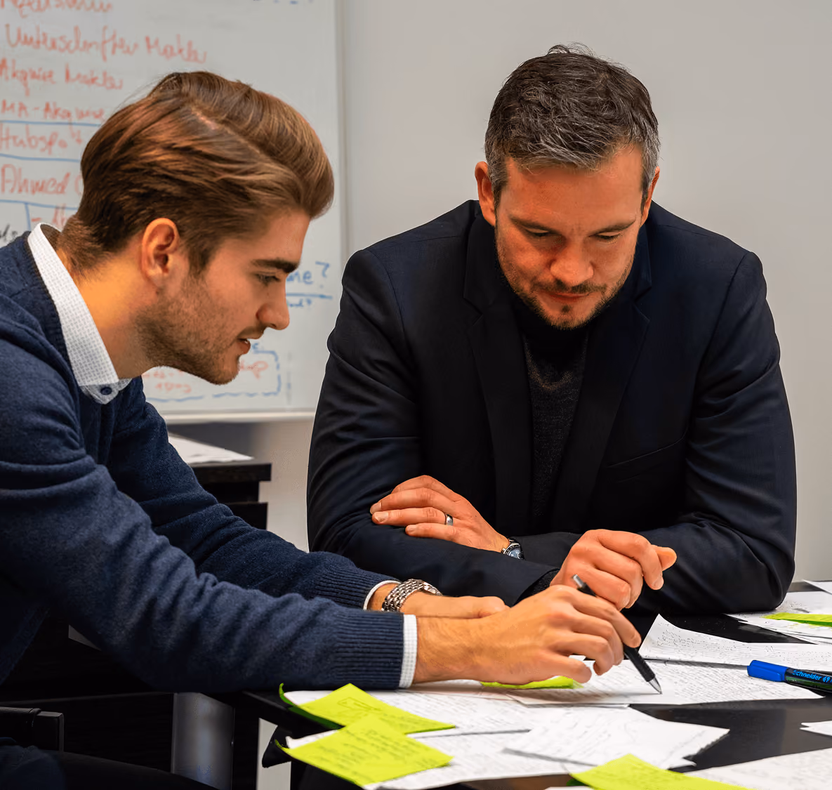 Zwei Männer besprechen sich an einem Tisch mit Notizen und Stiften in einem Büro.