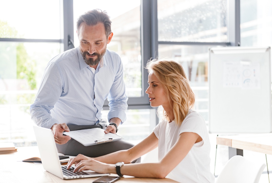 Ein bärtiger Mann, der einen Klemmbrett hält, lehnt sich über eine blonde Frau, die an einem Laptop arbeitet. Beide blicken konzentriert auf den Bildschirm in einem hellen, modernen Büro.