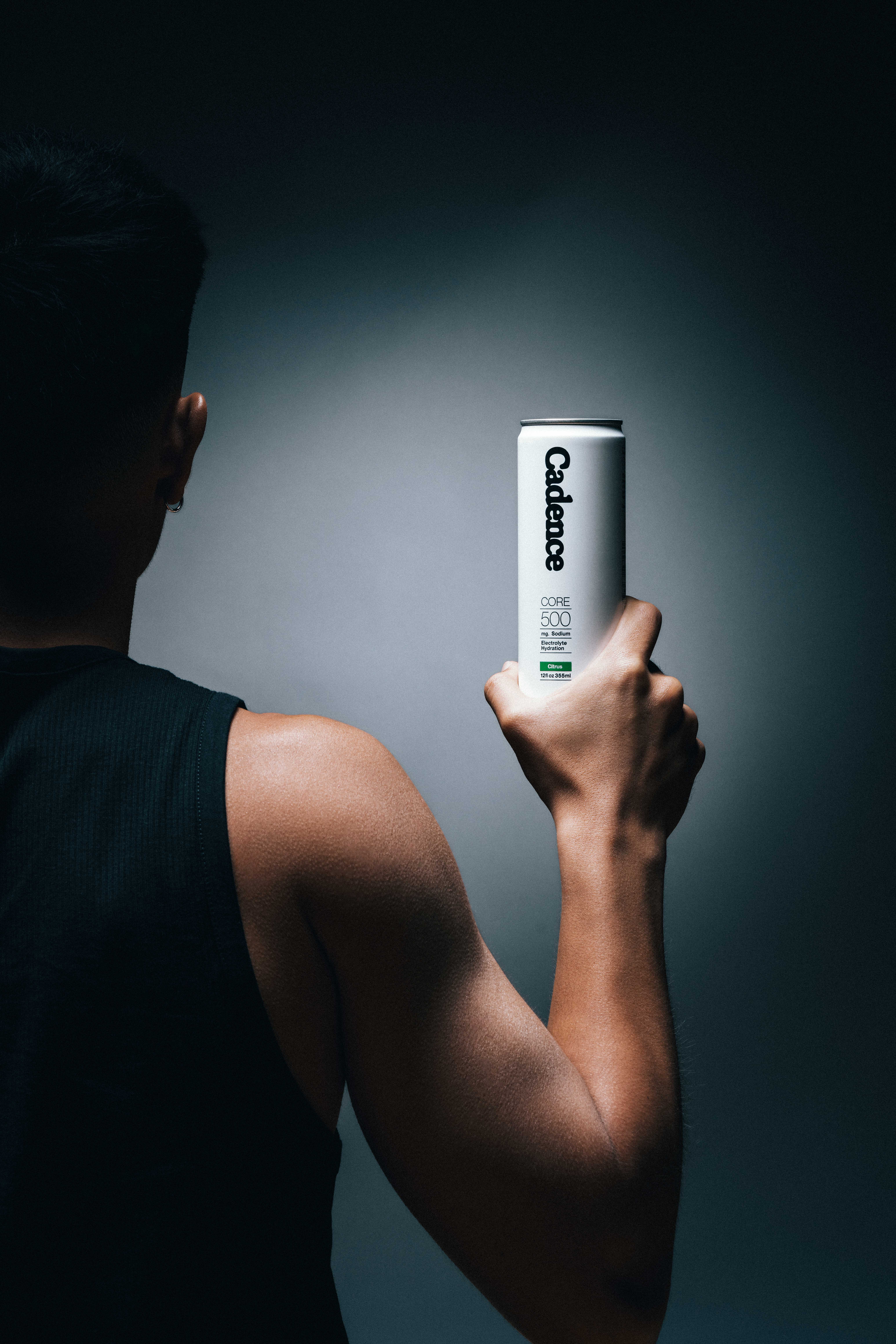 Back view of a person in a black sleeveless shirt holding a white can of Cadence electrolyte hydration drink.