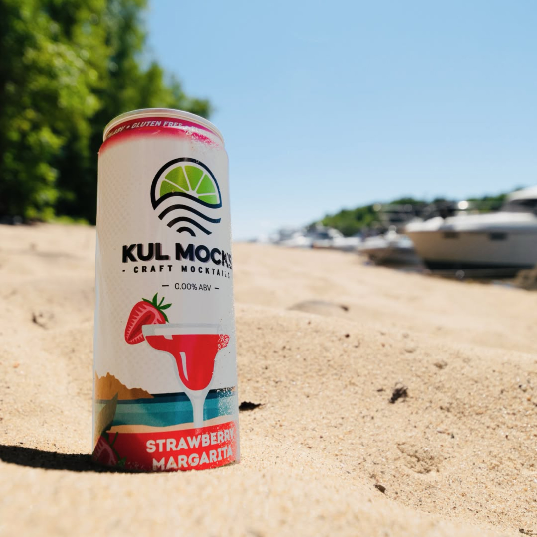 Can of Kul Mocks Strawberry Margarita craft mocktail on sandy beach with boats in background.