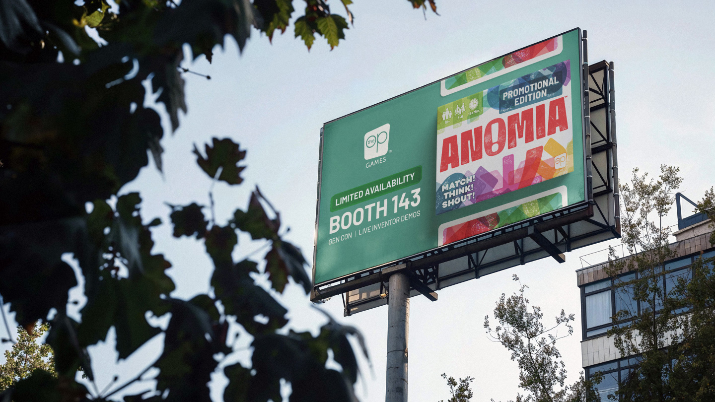 Billboard advertising Anomia game at Booth 143, limited availability, with trees and a building nearby.