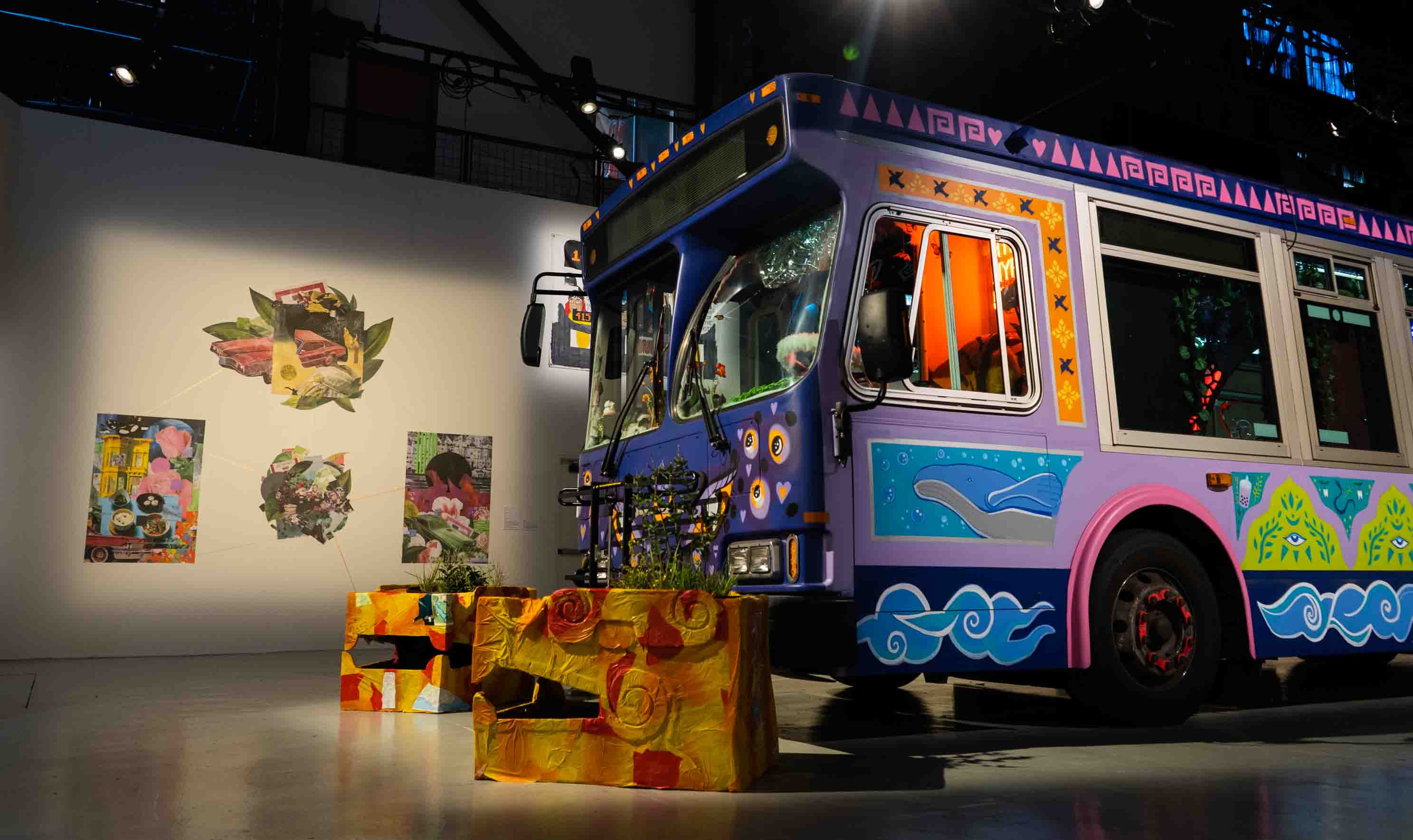 Colorfully painted purple bus with ocean and eye motifs displayed in an art gallery with illuminated wall collages and bright yellow planters.
