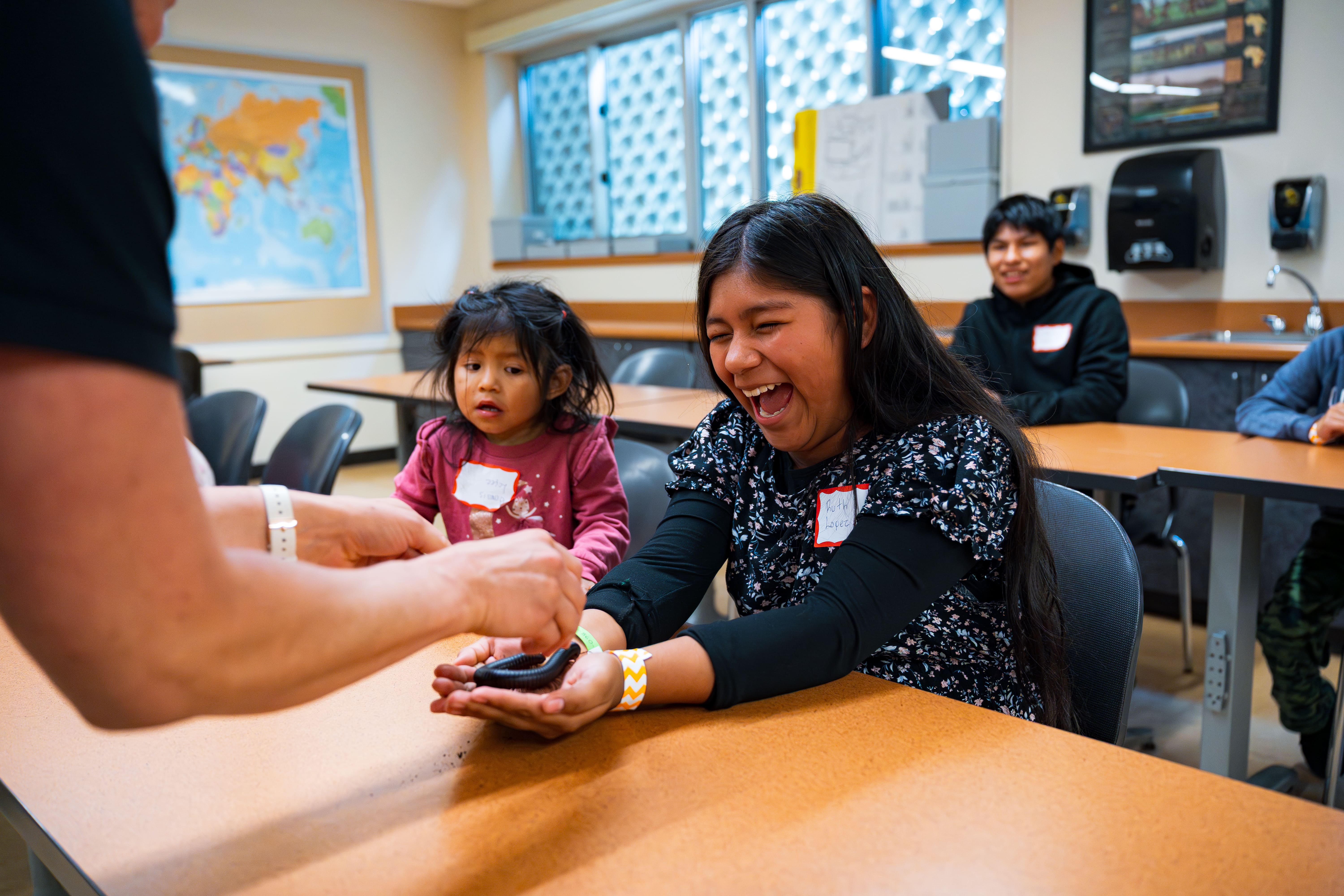 Smiling girl laughing while holding a large black millipede on her hands in a classroom setting.