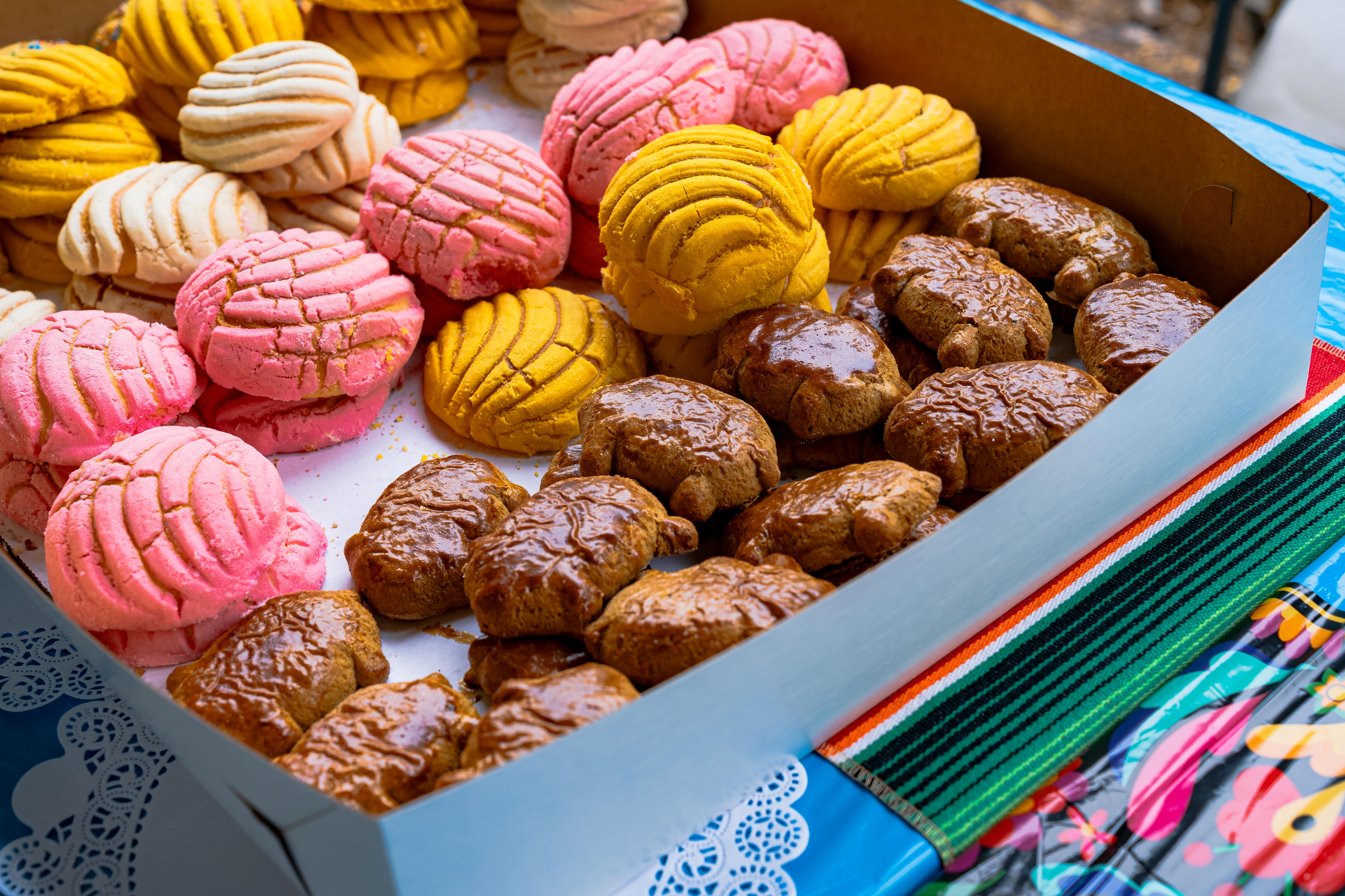 Box of colorful Mexican conchas and brown pastries on a striped cloth and a decorated tablecloth.