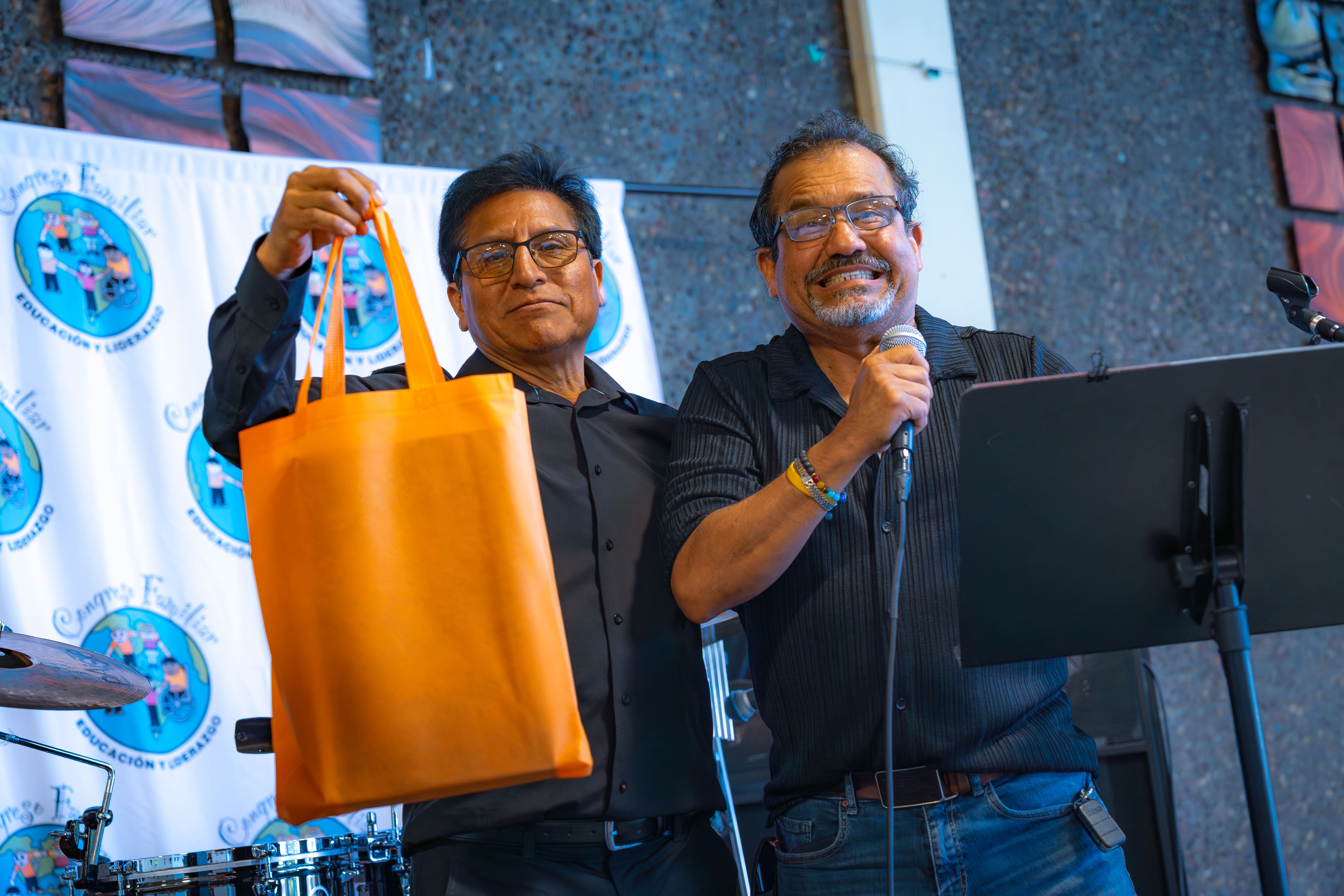Two men, one holding an orange tote bag and the other speaking into a microphone, standing in front of a backdrop with logos.