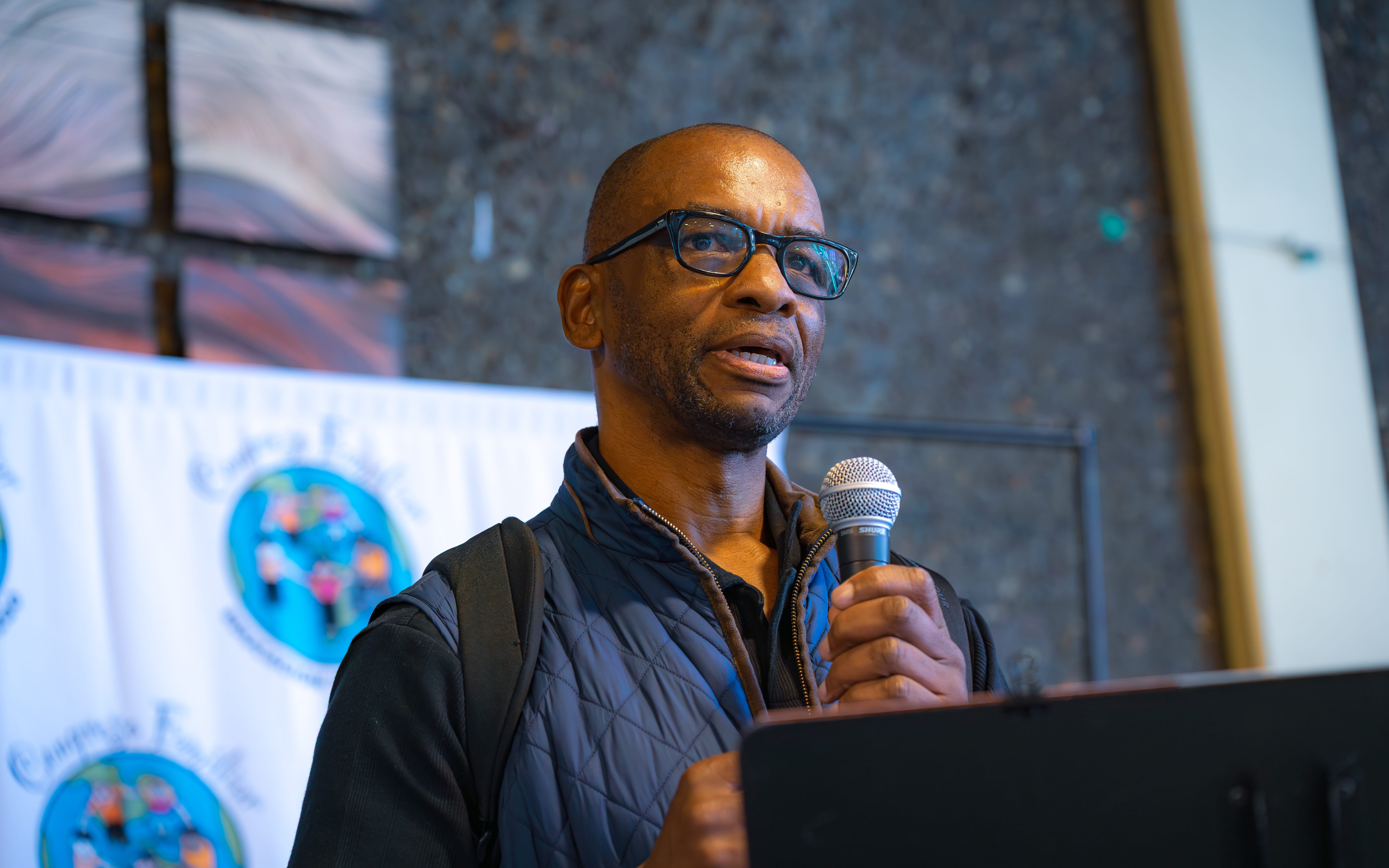 Man wearing glasses and blue quilted vest speaking into a microphone at a podium.