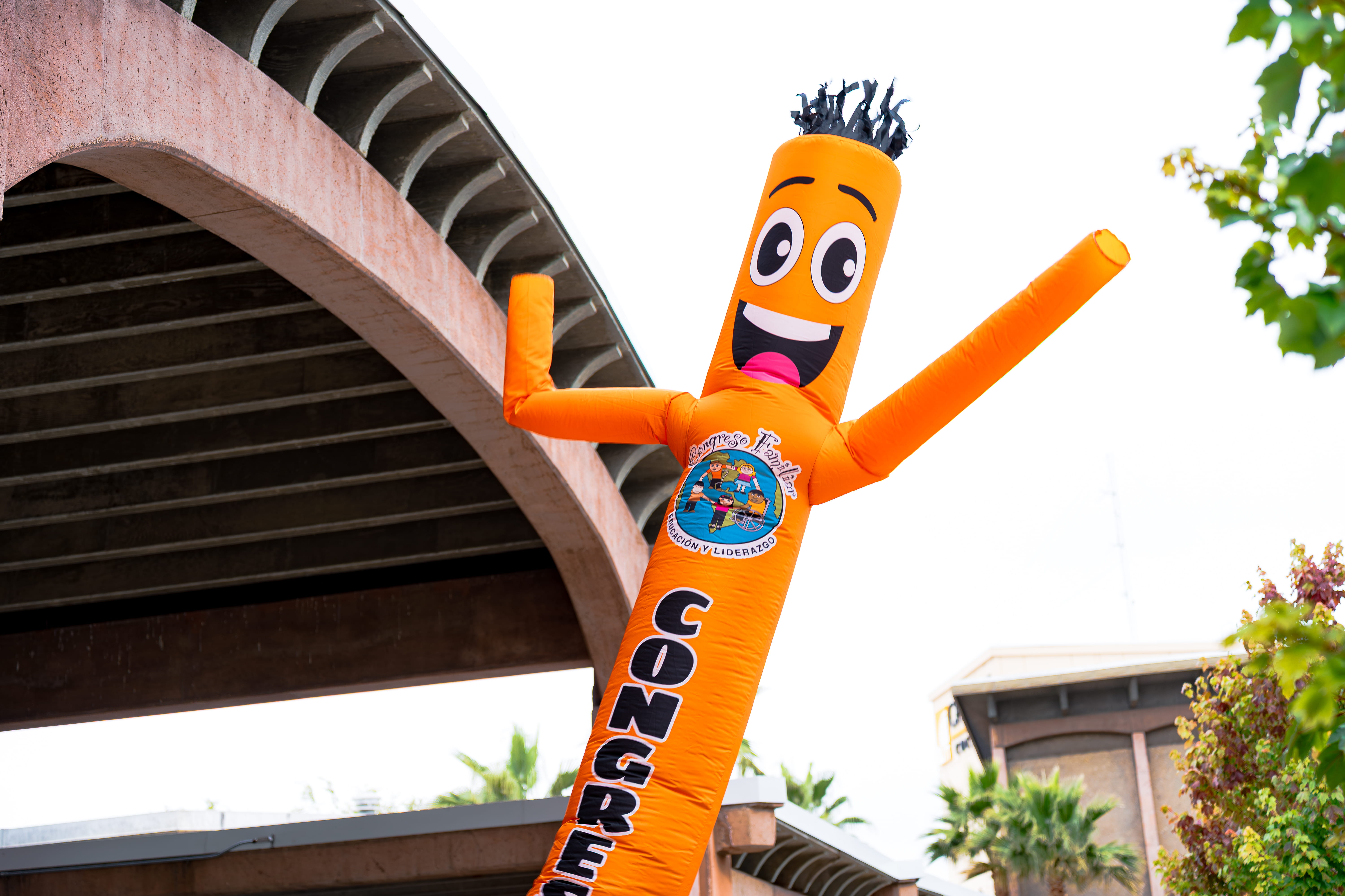 Orange inflatable tube man with a smiling face and black hair waving under a roof near trees and buildings.