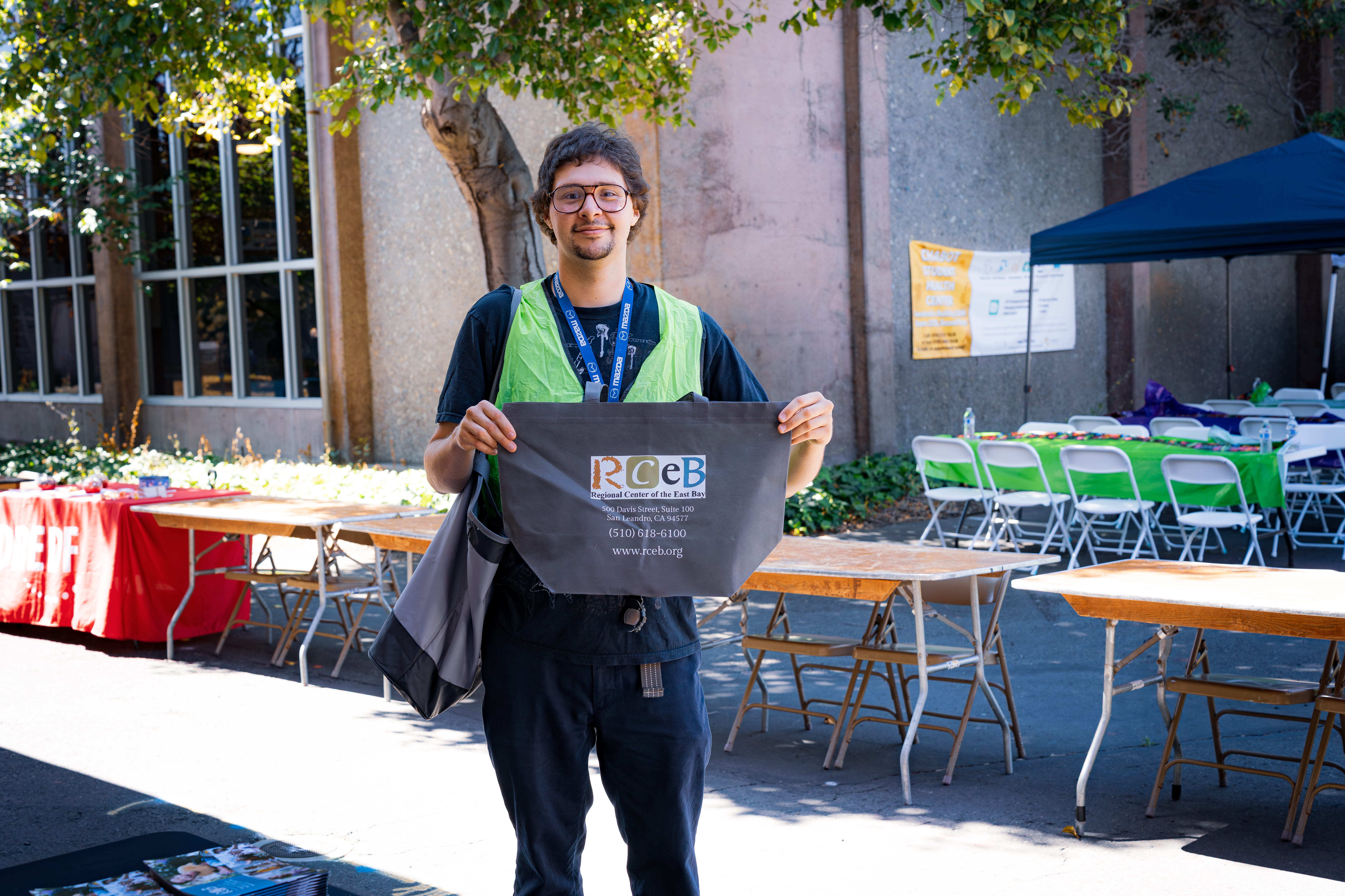 Man wearing glasses and a green safety vest holding a tote bag with RCEB logo at an outdoor event with empty tables and chairs.