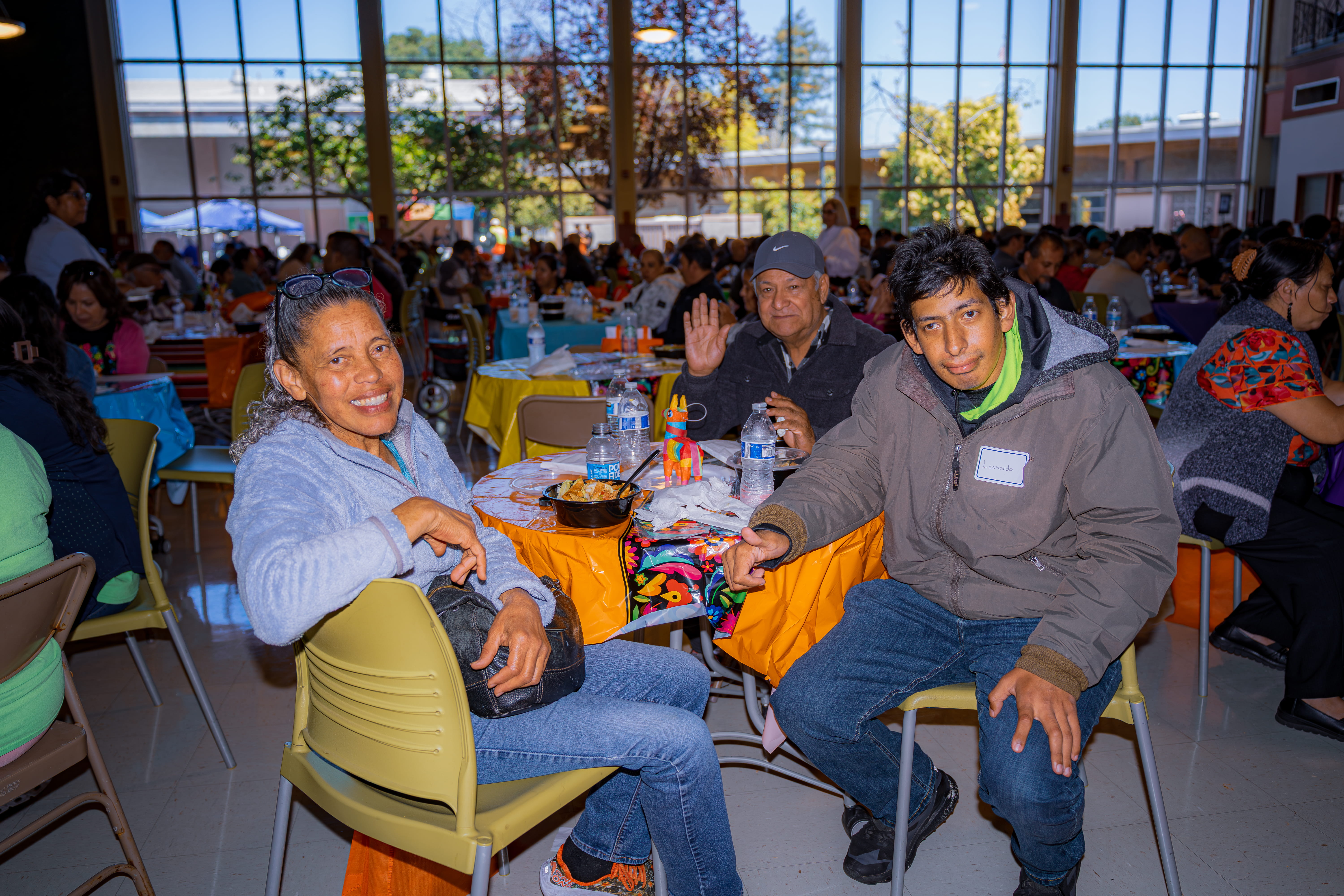 Three people seated at a table with food and drinks in a large bright room filled with many others dining.