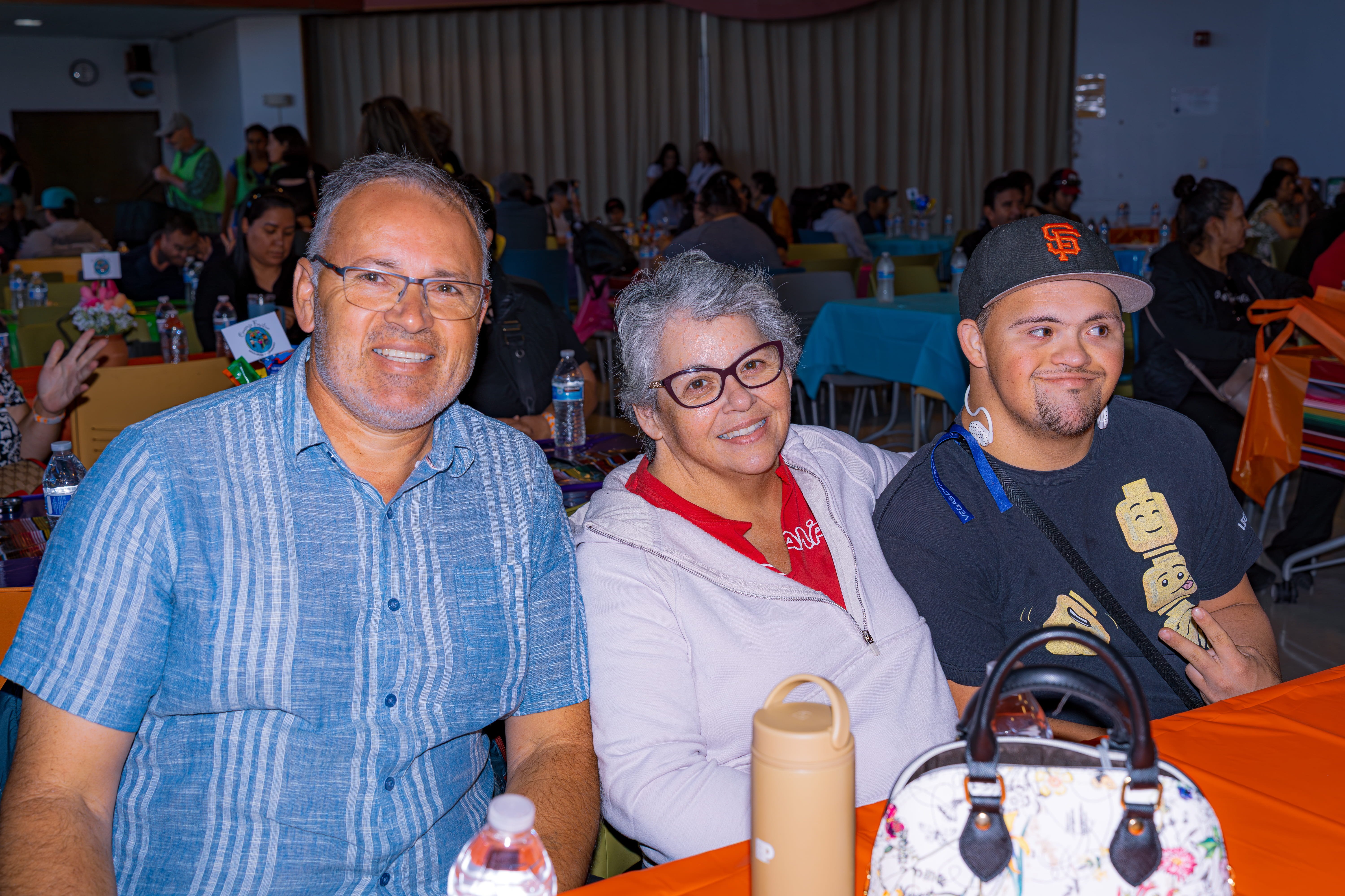 Three smiling people sitting at a table with orange tablecloth in a crowded event room.