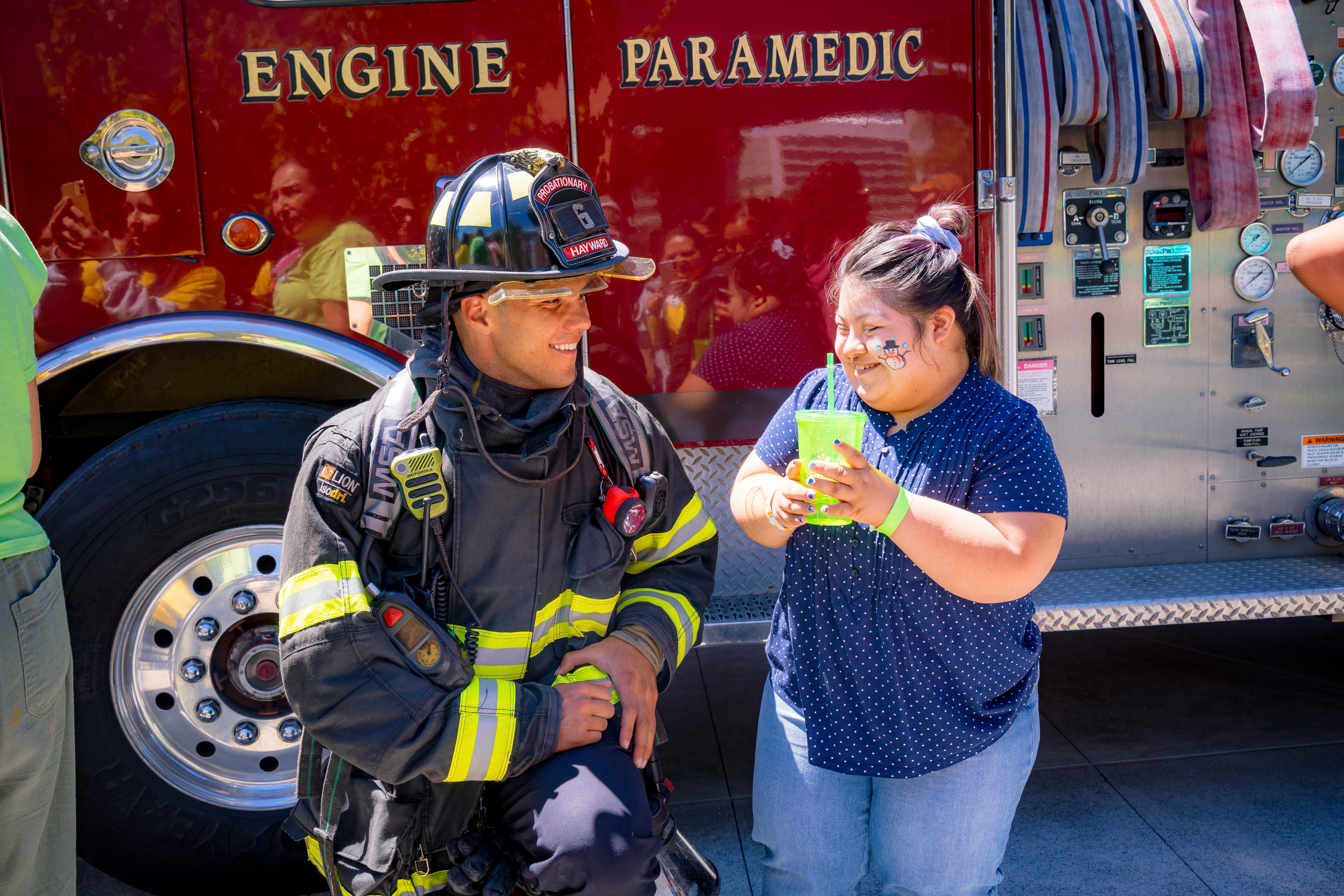Smiling firefighter in full gear kneeling next to a young girl with a snowman face painting, holding a green cup with straws in front of a red fire truck.